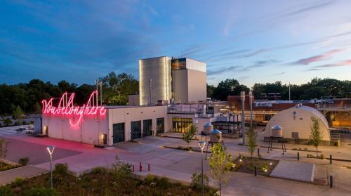 A modern building with a neon sign, surrounded by greenery and pathways, under a twilight sky.