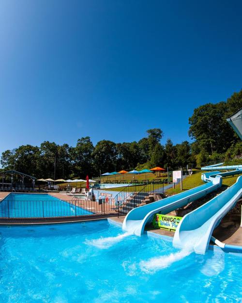 A sunny water park scene featuring blue water slides beside a swimming pool and colorful umbrellas in the background.