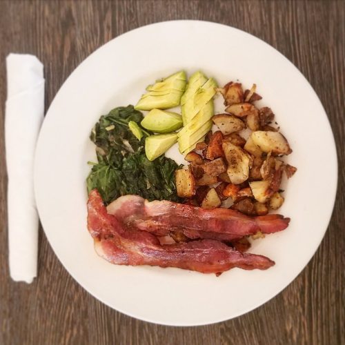 A plate with bacon, sautéed greens, avocado slices, and roasted potatoes on a wooden table.