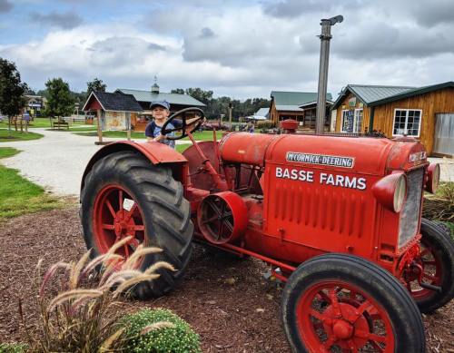 A child sits on a vintage red tractor at Basse Farms, surrounded by greenery and rustic buildings.
