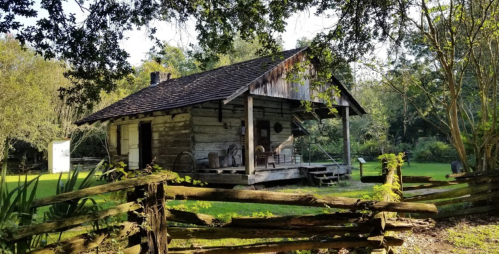 A rustic wooden cabin surrounded by trees and a wooden fence, with a porch and chairs in a serene outdoor setting.