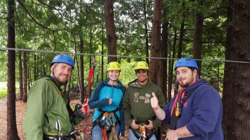 Four people in helmets and harnesses give thumbs up while standing on a zip line course in a forested area.