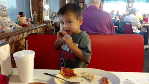A young boy in a gray shirt eats at a restaurant, with a plate of food and a drink beside him.