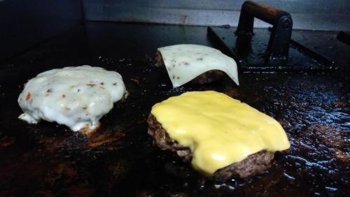Three burgers cooking on a griddle, two topped with melted cheese, one with a slice of white cheese.