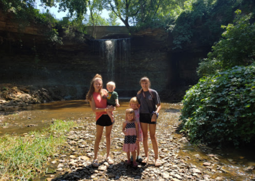 A group of four children stands by a rocky riverbank with a waterfall in the background, surrounded by greenery.
