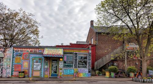 Colorful coffee shop with vibrant murals, surrounded by trees and a brick building, under a cloudy sky.