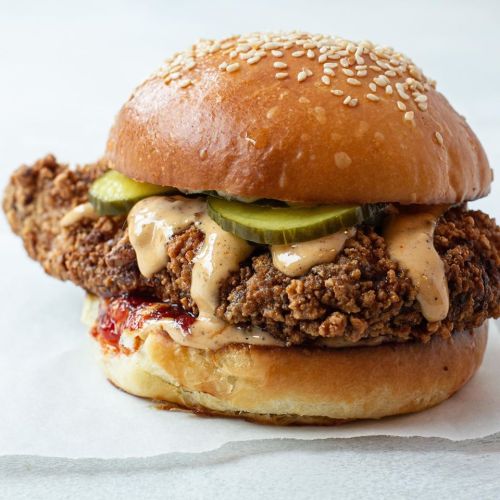 A crispy fried chicken sandwich with pickles and sauce on a sesame seed bun, served on parchment paper.