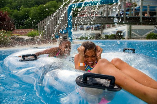 Two girls enjoy a water slide, splashing in a pool while one laughs and the other holds on tightly, both having fun.