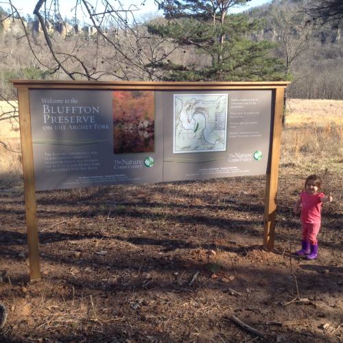 A child stands next to a sign welcoming visitors to Bluffton Preserve, surrounded by a natural landscape.
