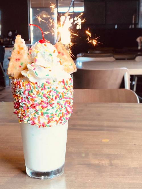 A colorful milkshake topped with whipped cream, sprinkles, a cherry, and a sparkler, served in a glass on a wooden table.