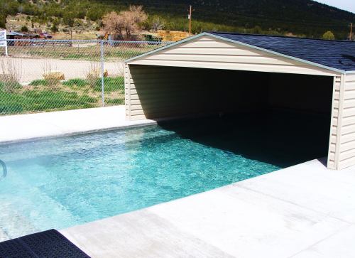 A covered swimming pool with clear water, surrounded by a concrete deck and a fenced area in a rural setting.