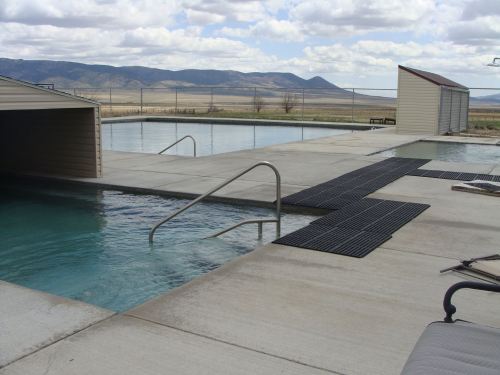 Two swimming pools with steps, surrounded by concrete and rubber mats, set against a backdrop of mountains and cloudy skies.