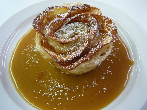 A dessert shaped like a rose, dusted with powdered sugar, sitting in a pool of caramel sauce on a white plate.