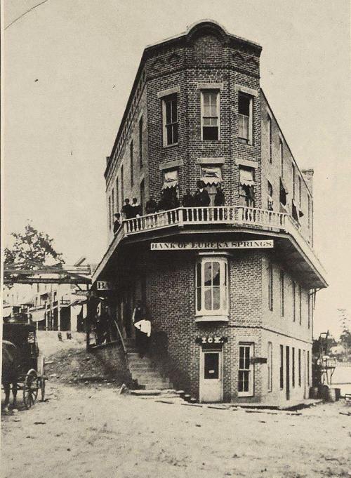 Historic brick building with a curved corner, labeled "Bank of Eureka Springs," featuring people on the balcony and steps.