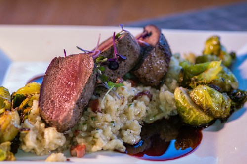 A plate featuring sliced beef, creamy risotto, and roasted Brussels sprouts, drizzled with a rich sauce.