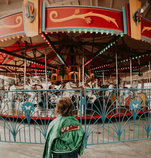 A child in a green jacket stands in front of a colorful carousel with illuminated horses and decorative fencing.