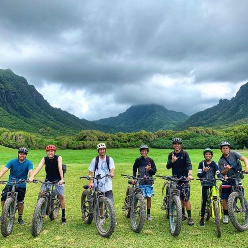 A group of eight people on mountain bikes, smiling in a lush green landscape with mountains and cloudy skies in the background.