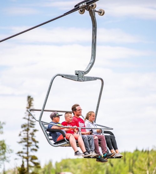 A group of four people riding a ski lift, surrounded by trees and a blue sky.
