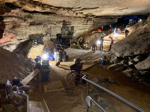 A dimly lit cave interior with workers, equipment, and lights illuminating the rocky surroundings and wooden structures.
