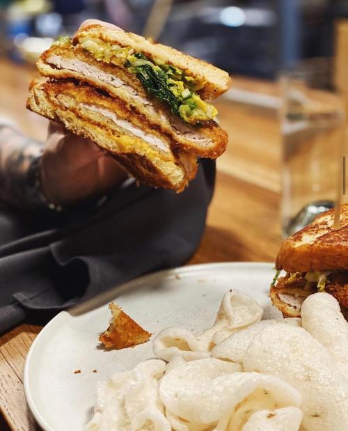 A hand holds a crispy sandwich filled with greens and meat, next to a plate of crispy snacks on a wooden table.