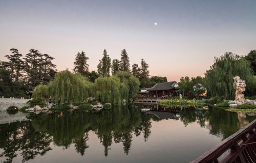 A serene garden scene at dusk, featuring a pond, lush trees, and a traditional building reflecting in the water.