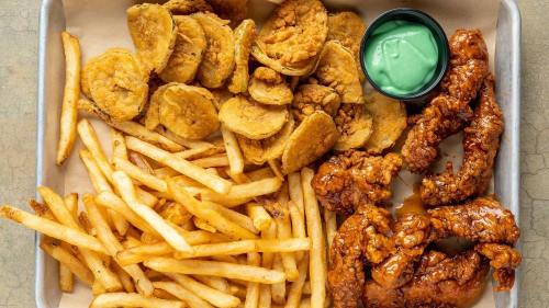 A tray of crispy fried chicken strips, golden fries, and crunchy fried pickles with a green dipping sauce.