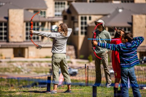 A group of people practicing archery outdoors, with one person aiming a bow and others observing in the background.
