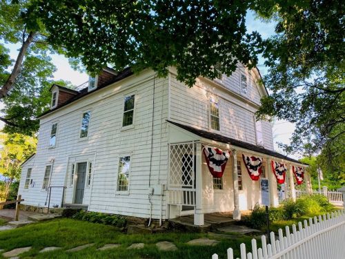 A white, two-story house with decorative red, white, and blue bunting, surrounded by greenery and a white picket fence.