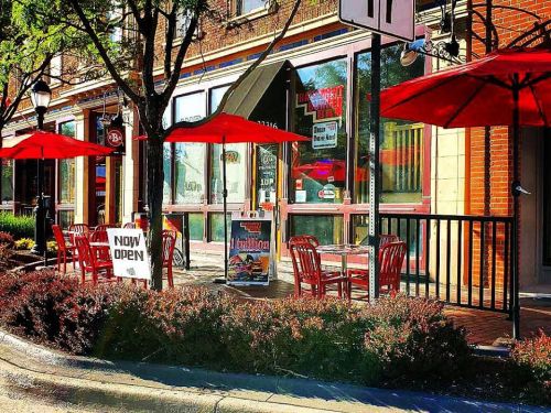 Outdoor seating area of a restaurant with red umbrellas, tables, and a "Now Open" sign on a sunny day.
