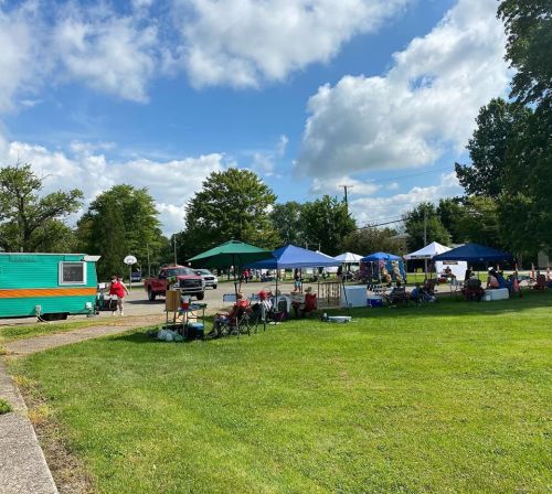 A vibrant outdoor market scene with tents, vendors, and people enjoying a sunny day on green grass.