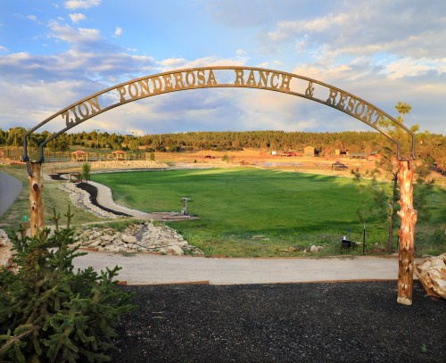 Entrance archway to Zion Ponderosa Ranch & Resort, with a scenic view of green fields and trees in the background.