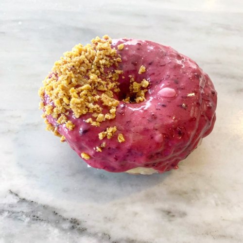 A glazed donut topped with purple berry icing and crumbled yellow topping, resting on a marble surface.
