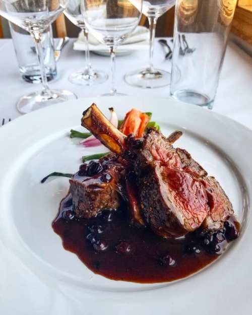 A plate of tender meat with a rich sauce, garnished with vegetables, set on a white tablecloth with wine glasses in the background.