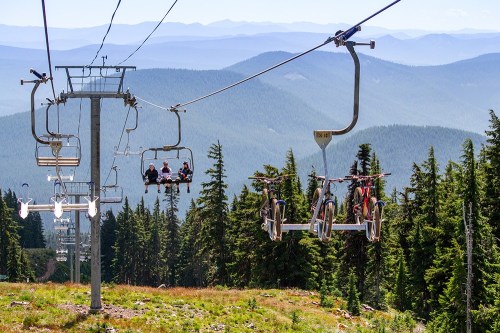 A scenic view of a ski lift with riders and bikes, surrounded by lush green trees and distant mountains.