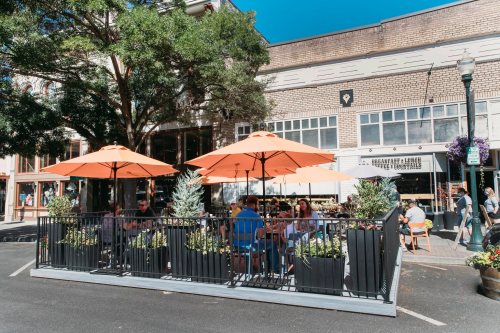 Outdoor dining area with orange umbrellas, surrounded by plants, bustling with people enjoying their meals.