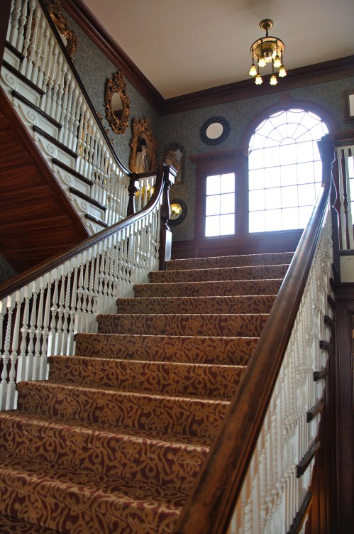 A grand staircase with ornate railings and patterned carpet, leading to a large window and decorative wall mirrors.