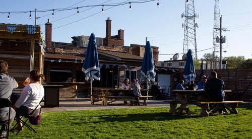 Outdoor seating area with people at picnic tables, string lights overhead, and a rustic bar in the background.