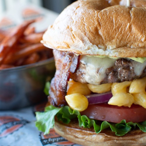 A close-up of a juicy burger with bacon, cheese, lettuce, tomato, and macaroni, served with a side of sweet potato fries.