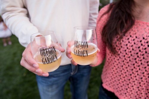 Two people hold glasses with "Oregon Garden" printed on them, filled with a light-colored beverage, outdoors on grass.