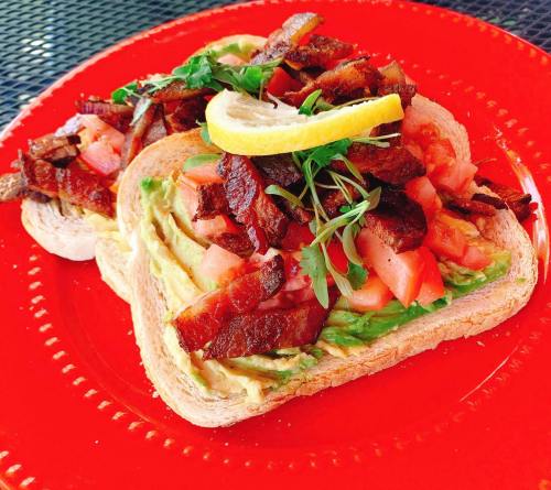 Two slices of toast topped with avocado, diced tomatoes, crispy bacon, and a lemon wedge, served on a red plate.