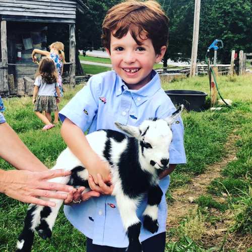 A smiling boy in a blue shirt holds a small black and white goat on a farm, surrounded by greenery and other children.