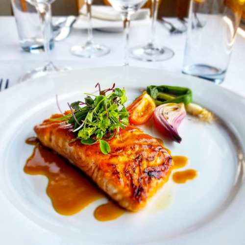 Grilled salmon fillet garnished with microgreens, served with colorful vegetables and a drizzle of sauce on a white plate.