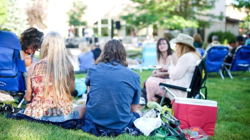 A group of people enjoying a picnic on the grass, seated in folding chairs, with a stage in the background.