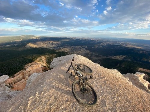 A mountain bike rests on a rocky ledge, overlooking a vast, scenic landscape with mountains and clouds.