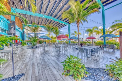 Outdoor dining area with palm trees, colorful umbrellas, and wooden flooring under a blue sky.