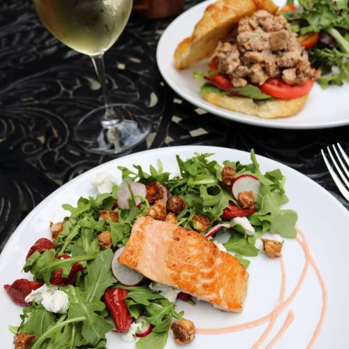 A plate with salmon on a bed of arugula, strawberries, and goat cheese, alongside a sandwich and a glass of white wine.