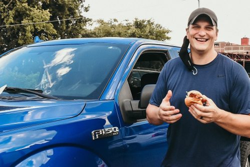 A smiling man holds a burger next to a blue truck, enjoying a sunny day outdoors.