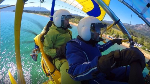 Two people in helmets flying in a yellow and blue ultralight aircraft over a clear blue lake and sandy beach.