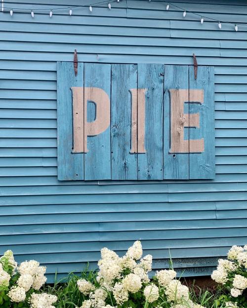 A blue wooden sign with the word "PIE" in large, light-colored letters, hanging on a blue wall, surrounded by white flowers.