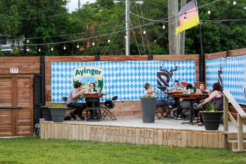 Outdoor beer garden with patrons at tables, decorated with blue and white checkered patterns and string lights.